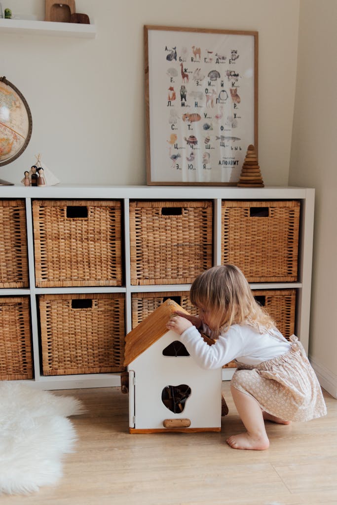 A cute toddler plays with a dollhouse in a cozy room with wooden storage baskets.
